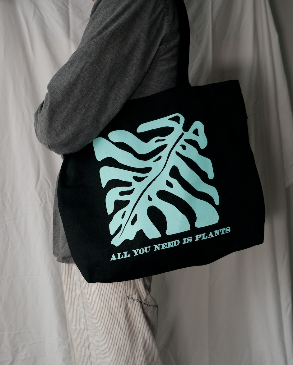 Black tote bag with a Monstera plant  leaf print and text held by a person against a neutral background
