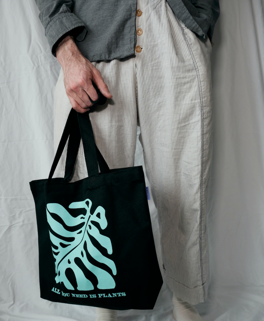 Person holding a black tote bag with a tropical plant green leaf design and text.